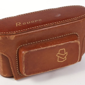 May include: A brown leather camera case with the word "Revere" embossed on the top and a gold logo on the front.