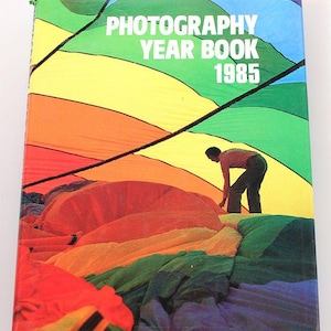 Puede incluir: Un libro de tapa dura titulado "Photography Year Book 1985" con una cubierta colorida. La cubierta presenta a una persona trabajando en una gran estructura de tela de colores del arcoíris. El lomo del libro es negro.