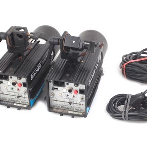 May include: Two black studio strobe lights with attached reflectors and adjustable arms. The lights have control panels with dials and buttons. Two black power cords are also included, one with a red band.