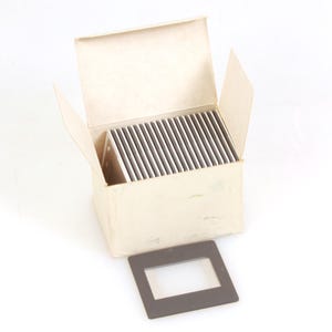 May include: A box of vintage 35mm slides, with a single slide frame in front. The box is open, revealing a stack of slides. The box and frame are a neutral color, set against a white background.