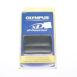 Puede incluir: Una cartera para tarjetas xD-Picture Card Olympus en un embalaje de plástico transparente. La cartera negra es rectangular, con cierre de solapa. El embalaje es azul y amarillo, con el logotipo de Olympus y el texto "xD-Picture Card".