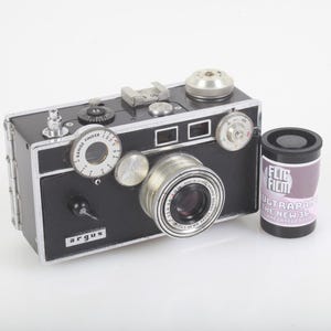 May include: A vintage black Argus camera with silver accents and a lens. A roll of Flic Film, labeled "ULTRA PAN THE NEW 36" is next to the camera. The camera has a range finder and various dials.