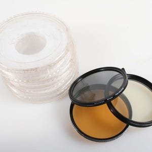May include: A stack of clear, circular lens protectors and three camera lens filters. The filters include a gray, a yellow, and a clear filter with black rims. The filters are labeled "Polarizing" and "67mm".