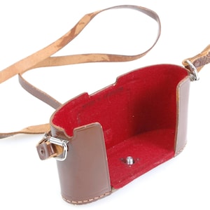 May include: A brown leather camera case with a red interior and a matching leather strap. The case is open, revealing the red lining. The strap is attached to the case with metal hardware.