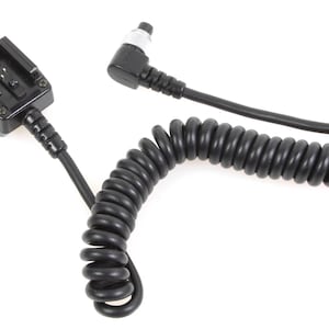 May include: Black camera flash sync cord with a coiled section and a hot shoe mount on one end. The other end has a right-angle connector. The cord is designed to connect a flash unit to a camera.