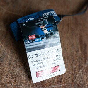 May include: A blue wrist strap with the text "GOTCHA WRIST STRAP" and "OP/CH USA" printed on a white label. The label also includes the text "Secures cameras, videos or binoculars comfortably around your wrist!" and "Made 100% in the U.S.A."
