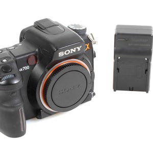 Puede incluir: Cámara réflex digital Sony α700 negra con tapa de objetivo y un cargador de batería negro separado. La cámara tiene un anillo naranja alrededor de la montura del objetivo.