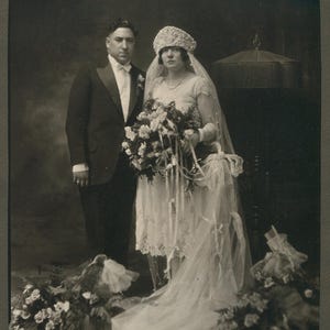 May include: Vintage black and white photograph of a couple on their wedding day. The bride wears a lace dress, veil, and beaded headpiece, holding a bouquet. The groom is in a tuxedo. Floral arrangements are at their feet, and a decorative object is in the background.