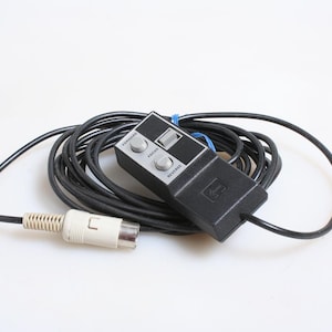 Puede incluir: Un mando a distancia de cámara negro y gris con un cable negro enrollado y un conector blanco. El mando tiene botones etiquetados como "FORWARD", "FOCUS" y "REVERSE". Una goma elástica azul asegura el cable.