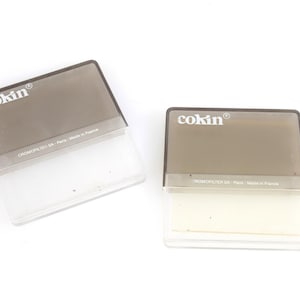 Puede incluir: Dos filtros de cámara rectangulares Cokin con diseño degradado. Cada filtro presenta una sección superior tintada en gris oscuro y una sección inferior transparente. La marca Cokin está impresa en blanco, junto al texto Promofilter SA Paris.