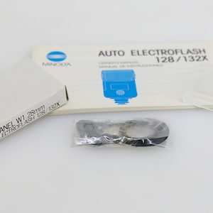 May include: A white box with the text "WIDE PANEL W1 28mm AUTO ELECTROFLASH 128/132X" printed on it. A white instruction manual with the text "AUTO ELECTROFLASH 128/132X" printed on it. A clear plastic wide panel for a flash unit. A small plastic bag containing a black ring.