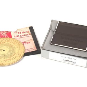 May include: A collection of vintage photography equipment, including a circular cine calculator, a film calculator, and a Schneider filter in a gray pouch. The calculator has a cream-colored face with black and red markings.