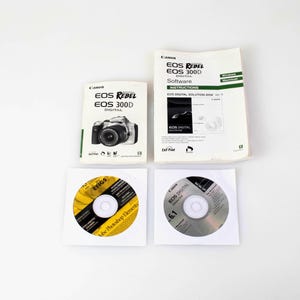 May include: A Canon EOS Rebel EOS 300D digital camera manual, a software instruction booklet, and two software discs in white paper sleeves. The manual features a camera illustration, while the software disc labels include text and branding.