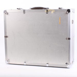 May include: A silver-colored, rectangular briefcase with a textured surface. The case has a brown handle and metal corner protectors. The case is closed and has metal latches and hinges. The background is white.
