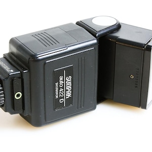 Może przedstawiać: Czarna lampa błyskowa do aparatu Sunpak auto 422 D Thyristor. Lampa ma prostokątny kształt z obrotową głowicą i białym reflektorem. Logo Sunpak i informacje o modelu są wydrukowane z boku.