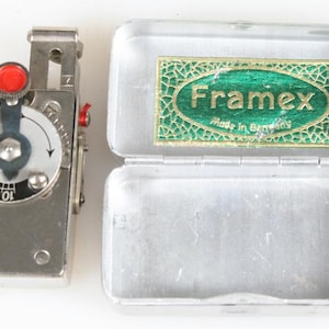 Peut inclure: Un posemètre Framex vintage et son étui en métal. Le posemètre est argenté avec un cadran noir et des accents rouges. L'étui ouvert porte une étiquette verte et dorée avec l'inscription "Framex Made in Germany."