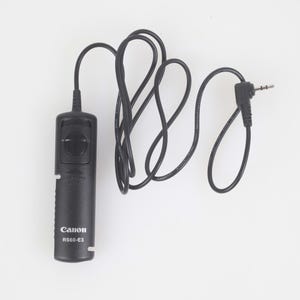 May include: A black Canon RS60-E3 remote shutter release with a coiled black cable and a 3.5mm jack. The remote has a button for controlling the camera's shutter. The Canon logo and model number are printed on the device.