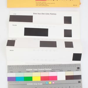 Puede incluir: Un conjunto de guías de separación de color y escala de grises Kodak con un sobre amarillo con texto, hojas blancas con parches rectangulares negros etiquetados como Cian, Amarillo, Magenta y Negro, y una tarjeta de control de 35,6 cm.