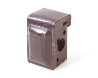 May include: A brown leather camera case with a stitched edge and a cut-out for the lens.