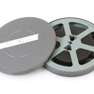 Puede incluir: Se muestra una bobina de película vintage y su estuche. La bobina es azul claro con la palabra "CARTOON" impresa. El estuche es gris con una etiqueta blanca con texto escrito a mano. La película es marrón oscuro.