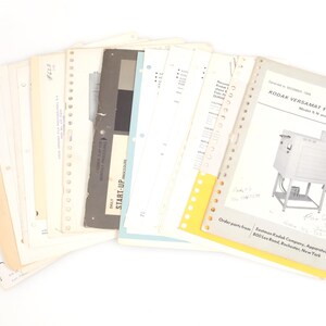 May include: A collection of vintage technical documents and manuals for a Kodak Versamat Film Processor, featuring white, beige, and grey paper sheets with black text, diagrams, and punched holes for binder storage, arranged in a fanned-out display.