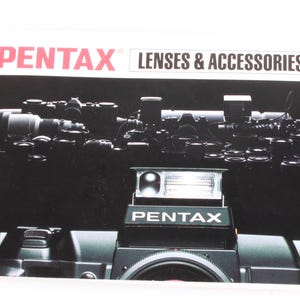 May include: A black and silver Pentax camera with a flash attached. The camera is on a black background with the text "PENTAX LENSES & ACCESSORIES" above it.
