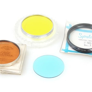 May include: Four camera filters in plastic cases. A yellow filter, a blue filter, an orange filter with the text "Kodak Wratten" and a black filter with the text "Spinatone Custom Crostar 2 SQ Set 7".