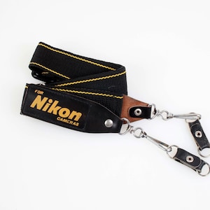 May include: Black and yellow Nikon camera strap. The strap has the Nikon logo in gold lettering. The strap is made of a black woven material with yellow stitching. The ends of the strap have metal clips and leather accents.