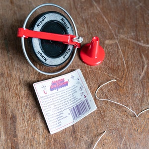 May include: A red and black spinning top toy with a white string. The top has the word "Nikon" printed on it four times. The instructions for the toy are printed on a white card with the words "Master the Forces!" printed in red and black.