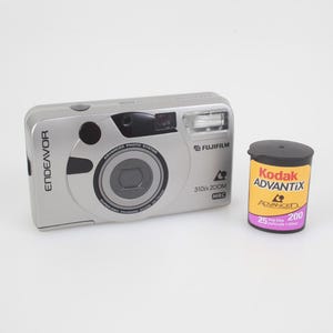 May include: A silver Fujifilm Endeavor 310 Zoom MRC camera with a black lens and flash, next to a Kodak Advantix film canister. The film canister is yellow, purple, and black.
