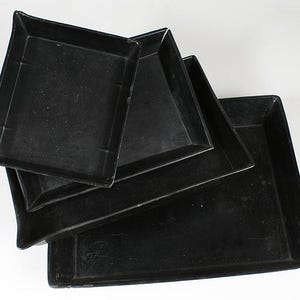 May include: A stack of four black square trays of varying sizes. The trays have slightly raised edges and a matte finish. The largest tray is rectangular, and the others are square. The trays are arranged in a nested configuration.