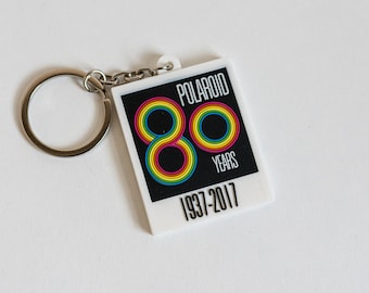 Puede incluir: Un llavero blanco con un logotipo Polaroid en blanco y negro y el texto "Polaroid 80 Years 1937-2017". El número 80 está hecho de círculos de colores arcoíris.