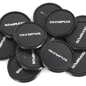 Op de afbeelding: Een verzameling zwarte Olympus lensdoppen, elk met de merknaam in witte letters. De ronde doppen, waarschijnlijk van plastic, hebben een matte afwerking. Ze liggen op een witte achtergrond.