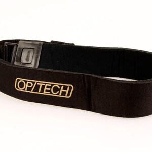 May include: Black camera strap with a metal buckle and the text "OP/TECH" in gold lettering. The strap is made of a soft, felt-like material and is designed to be worn around the neck or shoulder.