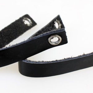 以下が含まれることがあります： 金属製のハトメが付いた黒い革のストラップが2本。 ストラップは、ざらざらした濃い色の内側と、滑らかで光沢のある外側をしています。 ハトメは銀色で、ストラップの端にあります。