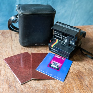 Peut inclure: Un appareil photo Polaroid Sun 600 LMS noir avec un étui de transport noir et un manuel d'instructions. L'appareil photo a une bande arc-en-ciel sur le devant et le texte "Polaroid" et "Sun 600 LMS" imprimé dessus.