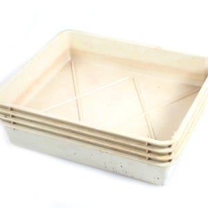 May include: A stack of four off-white, square plastic trays. The trays have a slightly textured surface and a shallow depth. The interior of each tray features a cross-shaped design for structural support. The trays are stacked on top of each other.