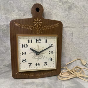 May include: A vintage General Electric wall clock with a wooden cutting board design. The clock face is square with black numbers and hands, and a floral design in the center. The clock is brown with decorative floral carvings.