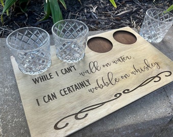 May include: A wooden whiskey serving tray with two recessed circles and engraved text that reads, "While I can't walk on water, I can certainly wobble on whiskey." Three diamond-patterned glasses are on the tray.