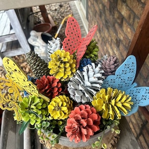 May include: A wooden planter with a rustic bark finish filled with painted pine cones, green foliage, and decorative butterflies. The pine cones are painted in shades of red, yellow, white, and blue.