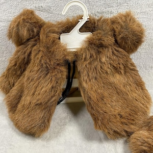 May include: A brown faux fur shrug featuring a thick, shaggy texture and rounded shoulder details. The garment is displayed on a white plastic clothing hanger, showcasing its open-front design and soft, dense synthetic fibers throughout the piece.