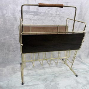 May include: A vintage magazine rack with a gold-colored metal frame and a dark brown wooden panel. The rack features a wooden handle and a wire grid design for storing magazines. The overall design is mid-century modern.