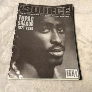 May include: A black and white magazine cover featuring Tupac Shakur. The magazine title "THE SOURCE" is at the top, with the text "THE MAGAZINE OF HIP-HOP MUSIC, CULTURE & POLITICS" below it. The cover also includes the text "TUPAC SHAKUR 1971-1996".