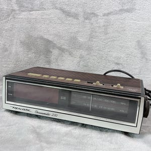 May include: A vintage Realistic Chronomatic-230 digital alarm clock. The clock has a brown top with several buttons, a digital display, and a white and silver face. The clock displays AM/FM radio frequencies.