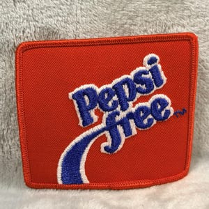May include: A square, red fabric patch with the Pepsi Free logo embroidered in blue and white. The patch has a stitched border and a curved blue accent beneath the text.