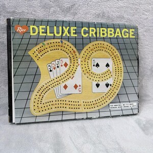 May include: A vintage Deluxe Cribbage game by Rico. The game board features a yellow track with card illustrations and the number 29. The box has a gray grid background with the words "DELUXE CRIBBAGE" in yellow.
