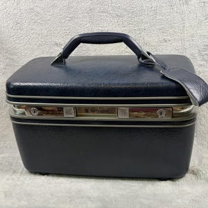May include: A dark navy blue hard-shell vanity case with a textured finish, a top handle, and a matching luggage tag. The front features a silver-toned metal trim with two integrated key locks and the Samsonite brand name printed in the center.