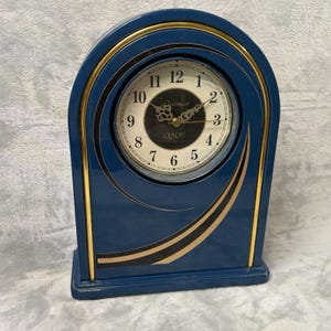 May include: A blue quartz clock with a gold trim and a white clock face. The clock has a rectangular base and an arched top. The clock face has black numbers and hands. The clock is labeled "Paramount Quartz".