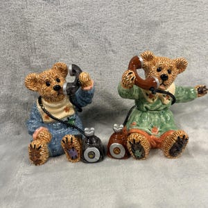 May include: Two ceramic bear figurines, each holding a telephone receiver. One bear wears a blue outfit, the other a green dress. Both have brown fur and are seated. The telephones are black and brown.