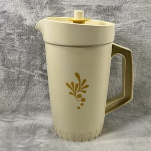 May include: A cream-colored plastic pitcher with a matching lid and handle. The pitcher features a gold floral design on the front. The lid has a yellow center. The pitcher is designed for holding liquids.